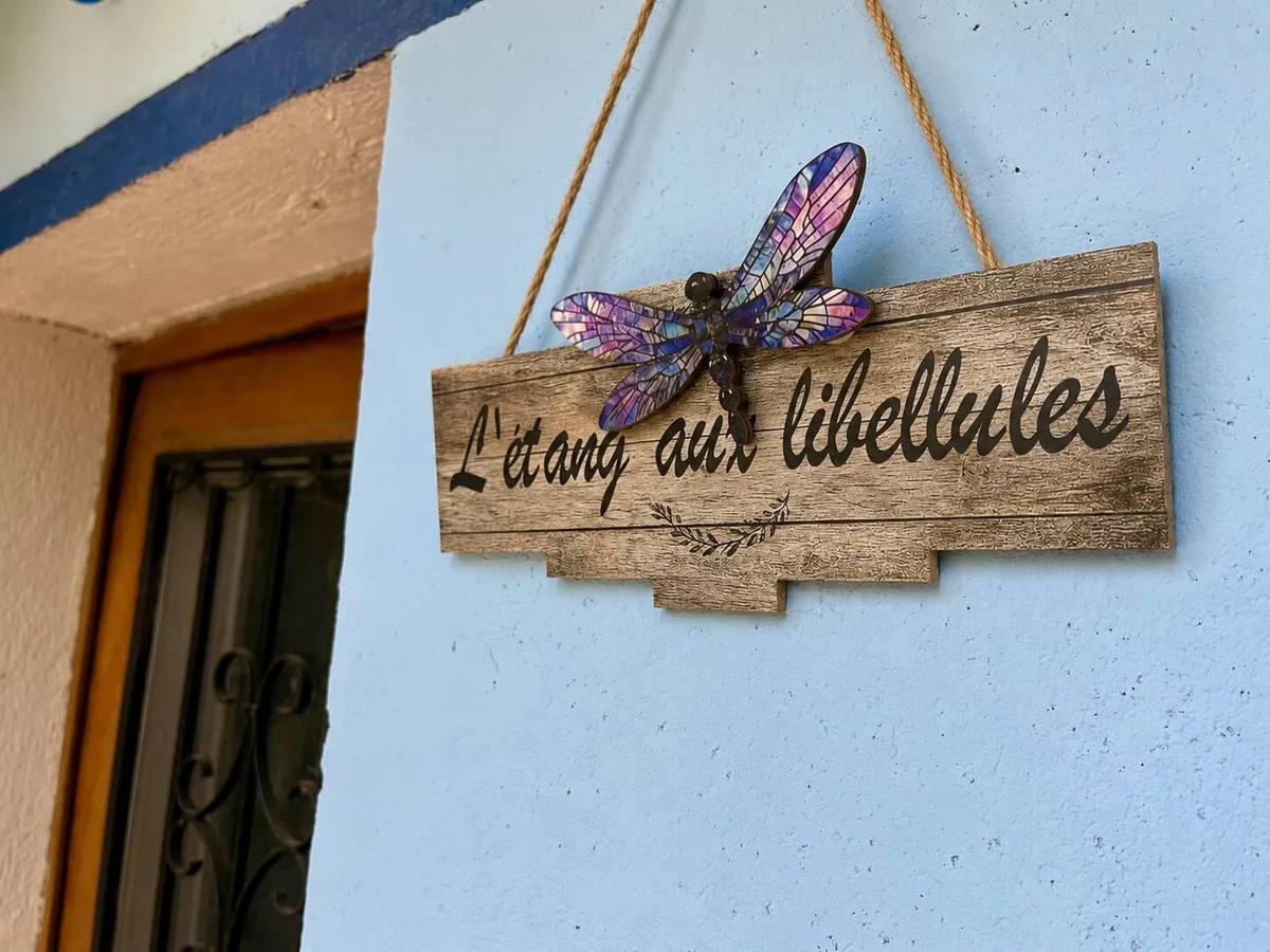 Le panneau en bois « L’étang aux libellules » orné d’une libellule, sur la façade bleue